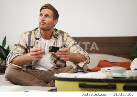 Portrait of young man booking tickets, going on holiday, holding credit card and smartphone, sitting with mobile phone near suitcase filled with clothes, ready for vacation trip Portrait of young man booking tickets, going on holiday, holding credit card and smartphone, sitting with mobile phone near suitcase filled with clothes, ready for vacation trip 113333103