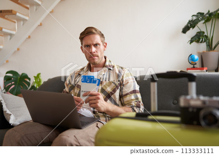 Portrait of serious man with laptop, holds two flight tickets, frowns and stares at camera with concerned, angry face expression Portrait of serious man with laptop, holds two flight tickets, frowns and stares at camera with concerned, angry face expression 113333111