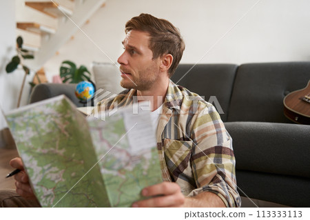 Portrait of young man, traveller, sitting packing suitcase, looking at travel map, choosing holiday destination, studying the route on vacation Portrait of young man, traveller, sitting packing suitcase, looking at travel map, choosing holiday destination, studying the route on vacation 113333113