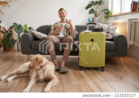 Image of excited man, tourist with a dog, sits in living room with packed suitcase and travel camera, guy saying yes and celebrating, pleased with prices online, booking pet-friendly hotel 113333117