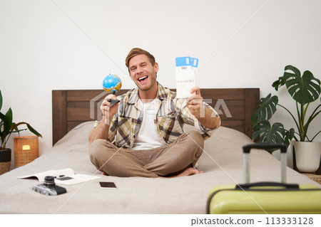 Portrait of happy guy, tourist goes on vacation abroad, holds globe and plane ticket, excited about his holiday, sits on bed with smiling, enthusiastic face expression 113333128