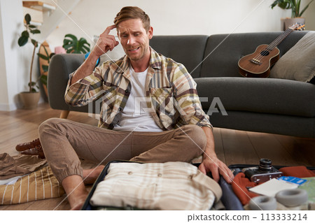 Portrait of worried man, tourist looking stressed and concerned, packing suitcase, holding hands on head, deciding on what to bring on holiday 113333214