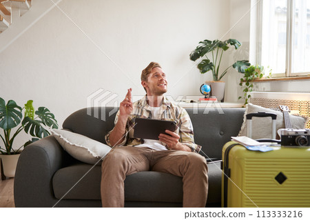 Excited young with tablet, sits in living room with packed suitcase, cross fingers for good luck, making wish, hoping for something Excited young with tablet, sits in living room with packed suitcase, cross fingers for good luck, making wish, hoping for something 113333216