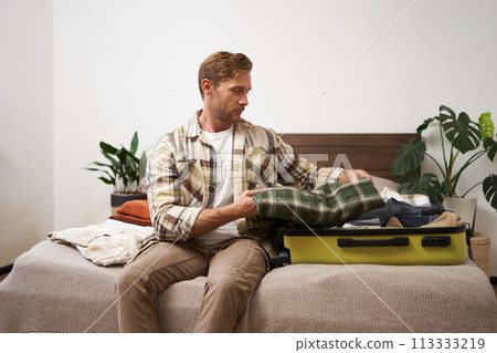 Portrait of young man packing his luggage for a trip, going on holiday, sitting on bed and putting clothes in suitcase, preparing for his flight Portrait of young man packing his luggage for a trip, going on holiday, sitting on bed and putting clothes in suitcase, preparing for his flight 113333219
