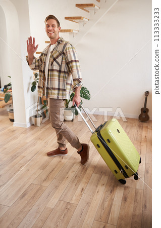 Vertical portrait of happy guy, tourist leaving the apartment with suitcase, waving hand at camera, saying hello or goodbye Vertical portrait of happy guy, tourist leaving the apartment with suitcase, waving hand at camera, saying hello or goodbye 113333233