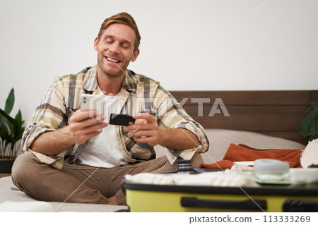 Portrait of young man, tourist paying online, using credit card and mobile phone app, sitting on bed with suitcase, ordering in internet, buying holiday tickets Portrait of young man, tourist paying online, using credit card and mobile phone app, sitting on bed with suitcase, ordering in internet, buying holiday tickets 113333269