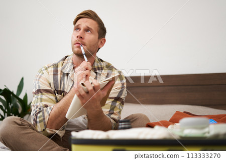 Portrait of thoughtful young man, tourist going on vacation, going through holiday check-list, writing down what he wants to take on travelling Portrait of thoughtful young man, tourist going on vacation, going through holiday check-list, writing down what he wants to take on travelling 113333270