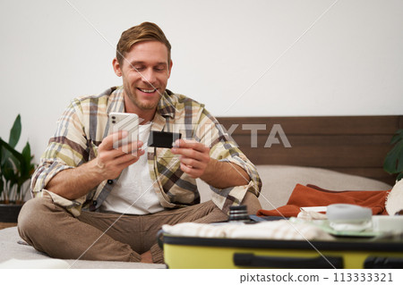 Portrait of young man booking tickets, going on holiday, holding credit card and smartphone, sitting with mobile phone near suitcase filled with clothes, ready for vacation trip Portrait of young man booking tickets, going on holiday, holding credit card and smartphone, sitting with mobile phone near suitcase filled with clothes, ready for vacation trip 113333321