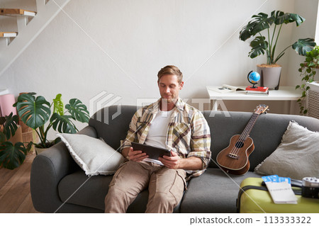 Portrait of young man in living room, sitting with tablet in hands, has suitcase packed and two plane tickets, preparing to go abroad, going on holiday 113333322