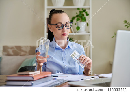Serious woman at workplace holding blister with capsules glass of water 113333511