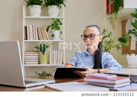 Business woman with clipboard sitting at workplace, looking talking at computer screen Business woman with clipboard sitting at workplace, looking talking at computer screen 113333512