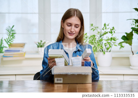 Young female customer sitting at home unpacking cardboard box with online purchases 113333535
