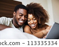 A man and woman are lying on a bed, smiling and looking at a tablet 113335334