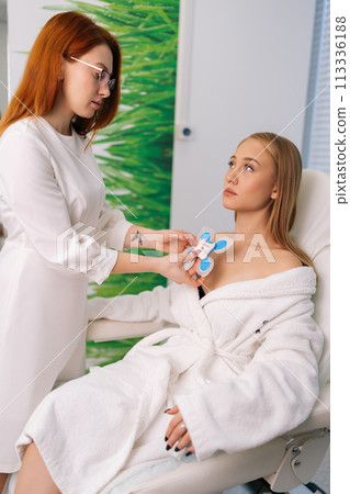 Vertical side view of skilled female cardiologist attaching holter sensors on woman patient with heart disease. Young woman during functional rehabilitation in cardiology room in clinic. 113336188