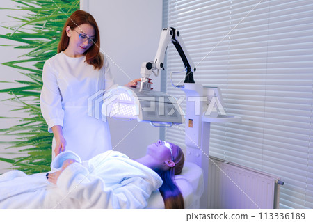 Portrait of pretty beautician specialist connecting and adjusting innovation device for skin rejuvenation at aesthetic clinic. Woman patient in protective goggles undergoes blue LED light therapy. Portrait of pretty beautician specialist connecting and adjusting innovation device for skin rejuvenation at aesthetic clinic. Woman patient in protective goggles undergoes blue LED light therapy. 113336189