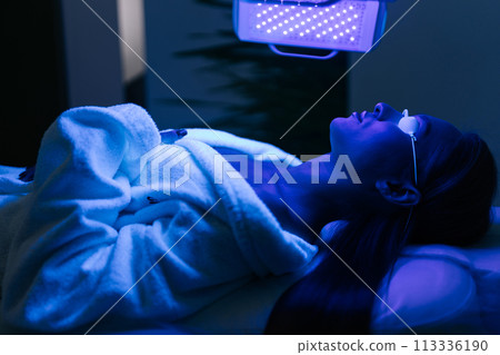 Young woman in glasses having blue LED light facial photodynamic therapy treatment in beauty salon. Female client having non-invasive type of phototherapy. Beauty, healthcare and wellness concept. Young woman in glasses having blue LED light facial photodynamic therapy treatment in beauty salon. Female client having non-invasive type of phototherapy. Beauty, healthcare and wellness concept. 113336190