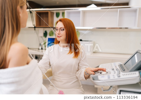Portrait of professional female doctor carrying out ultrasound examination breast of unrecognizable young woman patient in medical clinic. Diagnostician performing breast ultrasound procedure. 113336194