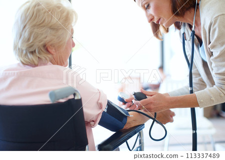 Ensuring her patients blood pressure is normal. Rear-view of a wheelchair-bound elderly patient getting her blood pressure checked by a nurse. Ensuring her patients blood pressure is normal. Rear-view of a wheelchair-bound elderly patient getting her blood pressure checked by a nurse. 113337489