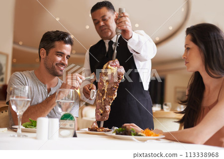 Couple, waiter and service for dinner in restaurant with meat, happiness and fine dining for anniversary or honeymoon. Man, woman and employee with steak on skewer, healthy meal and table in London 113338968