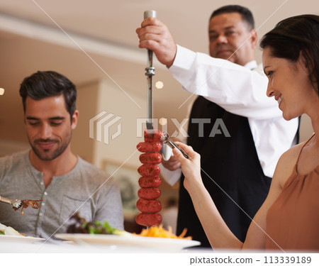 Couple, waiter and service for lunch in restaurant with meat, happiness and fine dining for anniversary or honeymoon. Man, woman and employee with sausage on skewer, healthy meal and table in London 113339189
