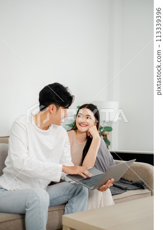 Young couple shares a joyful moment while looking at a laptop together on a cozy couch in a bright living room. Young couple shares a joyful moment while looking at a laptop together on a cozy couch in a bright living room. 113339396