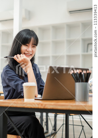 Cheerful young woman with pen in hand working on her laptop in a bright cafe, with a coffee cup on the table. 113339413