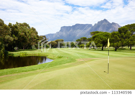 Hole, flag and grass at golf course in landscape of summer on luxury holiday or vacation. Calm, morning and lawn with turf for sport in woods, forest or nature of park with water, trees and field 113340596