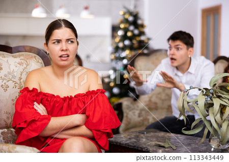 Displeased young European man and woman in red dress yelling at each other on sofa during preparations for New Year in living room 113343449