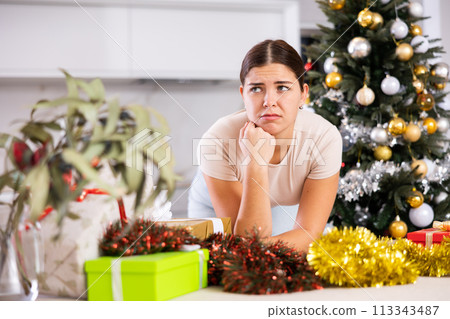 Emotional sad young female next to packed gift box with bow near and decorated Christmas tree at home 113343487
