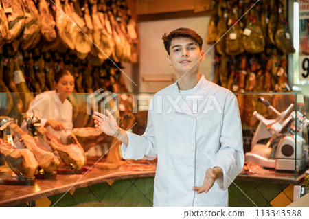 Young guy seller in white jacket selling spanish jamon in butcher shop Young guy seller in white jacket selling spanish jamon in butcher shop 113343588