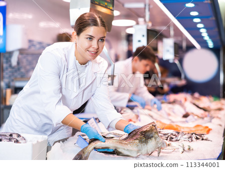 Positive saleswoman holding cod fish in fish store Positive saleswoman holding cod fish in fish store 113344001