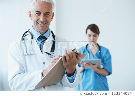 Portrait, mature doctor writing on his clipboard and with a nurse browsing on a digital tablet in the background. Senior doctor and nurse standing in corridor looking at files discussing reports 113344034