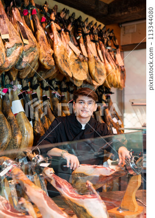 Male seller cutting slices from whole leg of jamon in meat store Male seller cutting slices from whole leg of jamon in meat store 113344080