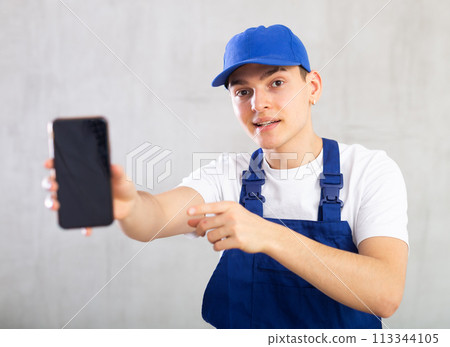 Man in jumpsuit and cap shows dark empty mobile phone screen and points at device with his hand 113344105