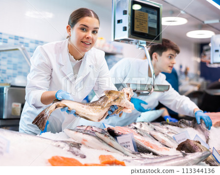 Positive saleswoman holding cod fish in fish store Positive saleswoman holding cod fish in fish store 113344136