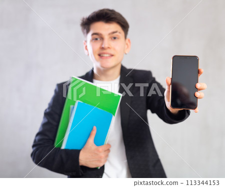 Guy in jacket with stack of notebooks shows empty mobile phone screen.Device close up unfocused 113344153
