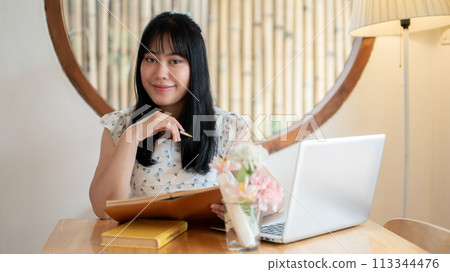A cute Asian woman sits at a table in a coffee shop with her laptop computer, smiling at the camera. A cute Asian woman sits at a table in a coffee shop with her laptop computer, smiling at the camera. 113344476