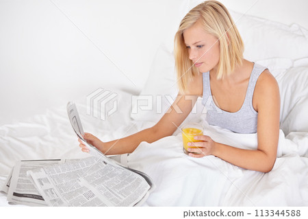 Young woman, orange juice and reading newspaper on bed for headlines, energy and health body with wellness. Blonde person, thirsty and glass of fruit drink to detox, morning and vitamin c in bedroom 113344588
