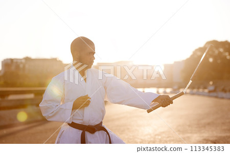 Man, karate and fitness with nunchuck in city for fighting style, technique or combat skill. Young male person or taekwondo fighter with weapon in martial arts or self defense on street in urban town 113345383