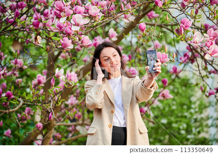 Happy woman taking selfie with smartphone among spring blossoms with soft focus on the background. 113350649