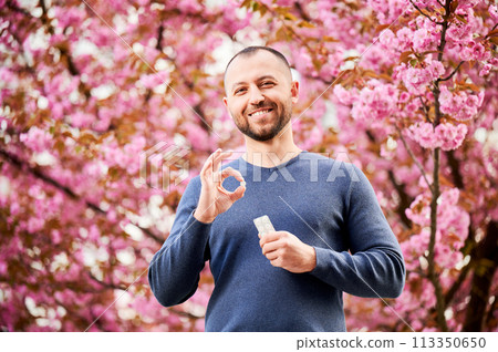 Man allergic suffering from seasonal allergy in blossoming garden at spring. Handsome smiling man holding allergy pill, making 'OK' hand sign in front of blooming tree. Spring allergy concept. 113350650