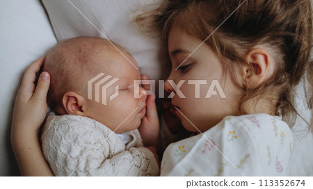 Portrait of big sister cuddling newborn, little baby. Girl lying with her new sibling in bed, closed eyes. Sisterly love, joy for new family member. Portrait of big sister cuddling newborn, little baby. Girl lying with her new sibling in bed, closed eyes. Sisterly love, joy for new family member. 113352674