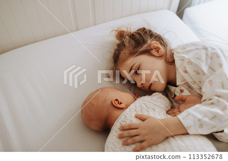 Portrait of big sister cuddling newborn, little baby. Girl lying with her new sibling in bed, closed eyes. Sisterly love, joy for new family member. Portrait of big sister cuddling newborn, little baby. Girl lying with her new sibling in bed, closed eyes. Sisterly love, joy for new family member. 113352678