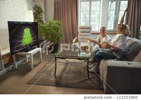 Two girls, friends sitting on couch at home and lively watching online football match, expressing positive emotions Two girls, friends sitting on couch at home and lively watching online football match, expressing positive emotions 113353960