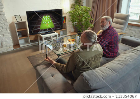 Two elderly men, sport fans sitting on couch at home in living room and watching online football match translation on tv. Supporting team 113354067