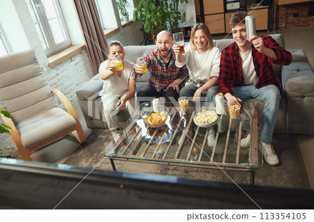 Young people, friends sitting in living room on sofa at home, eating snacks, drinking beer and watching online game translation 113354105