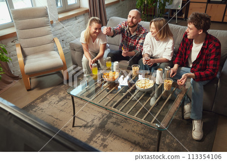 Young people, friends sitting in living room on sofa at home, eating snacks, drinking beer and watching online game translation 113354106