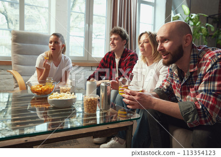 Tense game moment. Group of young people, friends gathering at home, sitting in living room, drinking beer and watching online match translation. 113354123