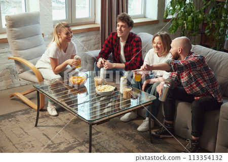 Happy young people, sport fans, men and women sitting on couch at home, eating snacks, drinking beer and watching online match translation Happy young people, sport fans, men and women sitting on couch at home, eating snacks, drinking beer and watching online match translation 113354132