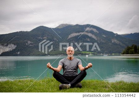 Senior man doing outdoor yoga, meditating by the lake in mountains. Elderly sportsman spending active vacation in nature. 113354442